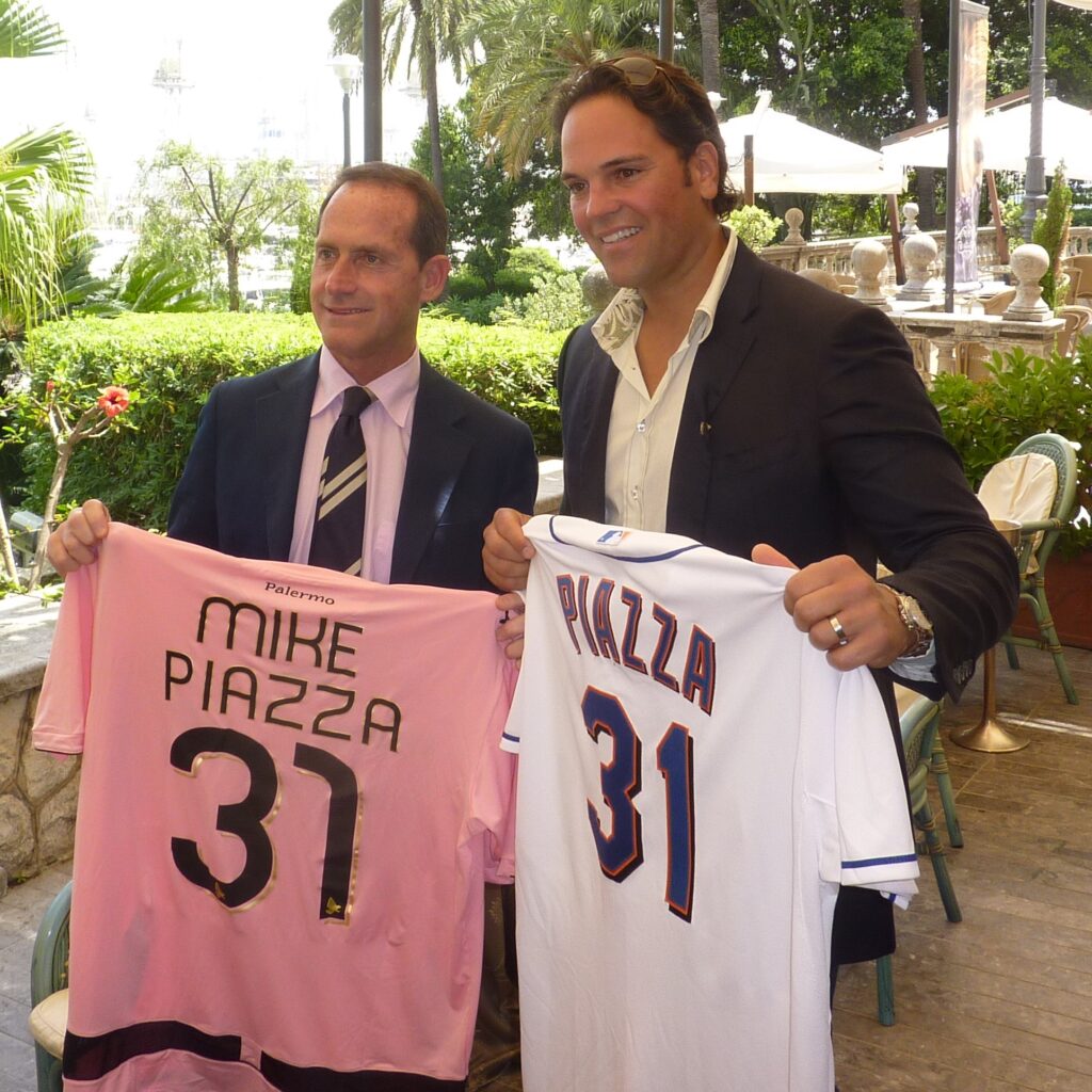 Mike PIazza holds up a Mets jersey with his name on it next to Palermo VP Micciche holding a Palermo jersey with Mike PIazza on it
